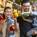 Happy husband and wife with a kid buys vegetables. Cheerful family of three choosing bell pepper and greens in vegetable department of supermarket or market.